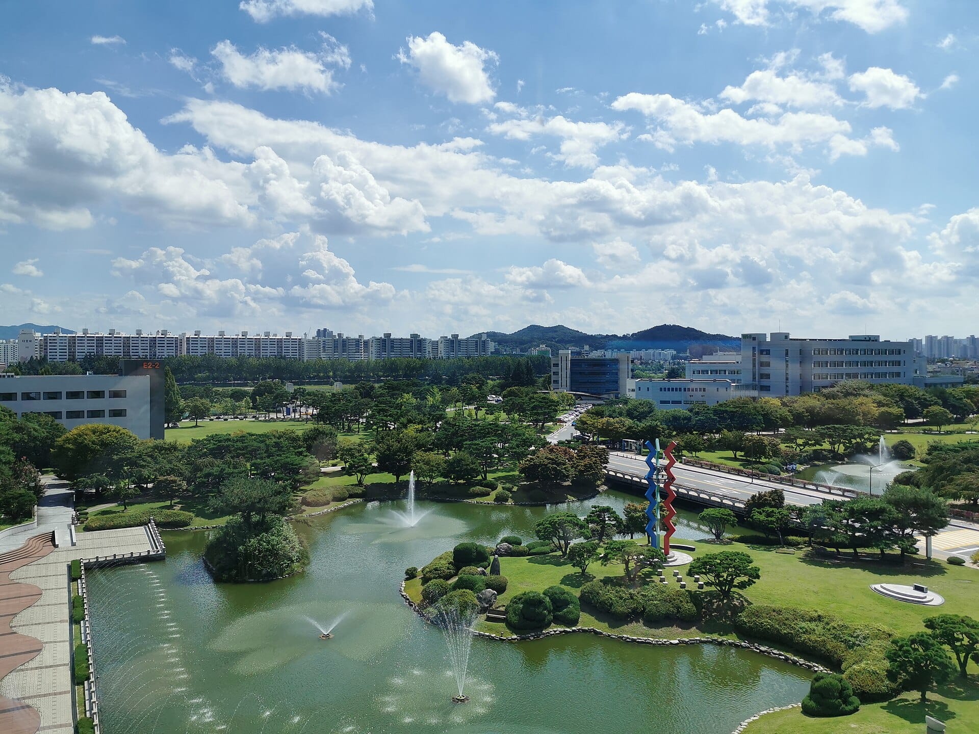 The KAIST campus in Daejeon with a landscaped pond and fountains under a cloudy blue sky